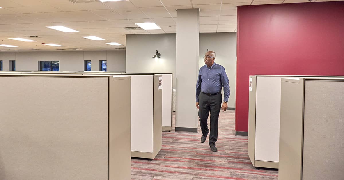 Office worker walking through cubicle aisle with red accent wall and overhead lighting in modern workplace
