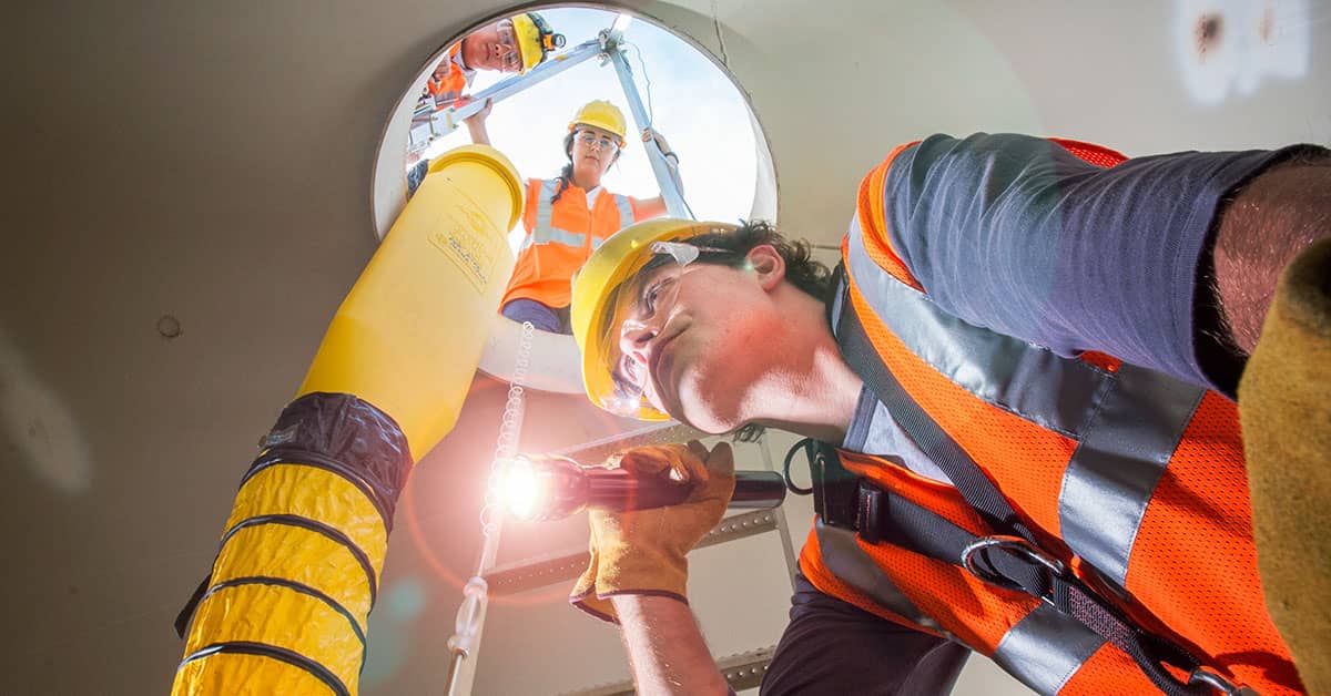 Construction workers inspecting a permit-required confined space with safety gear, flashlight, and ventilation hose, following OSHA safety protocols.