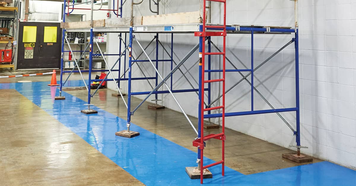 Indoor scaffold setup with metal frames and wooden platform, featuring red access ladder and safety base plates on a blue-painted floor.