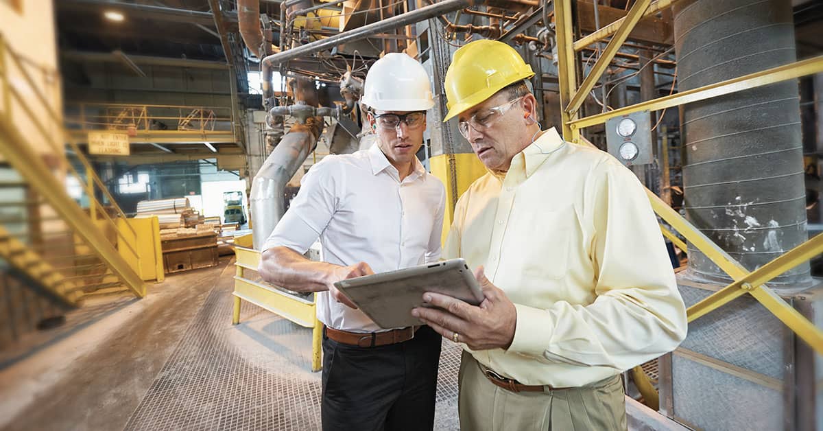 Two safety professionals wearing hard hats and safety gear reviewing plans on a tablet in an industrial factory setting with machinery and metal structures in the background.