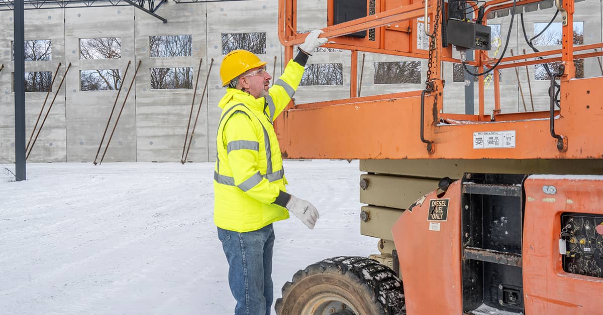 Worker in a high‑visibility winter jacket and hard hat inspecting an aerial lift on a snow‑covered construction site during freezing conditions.
