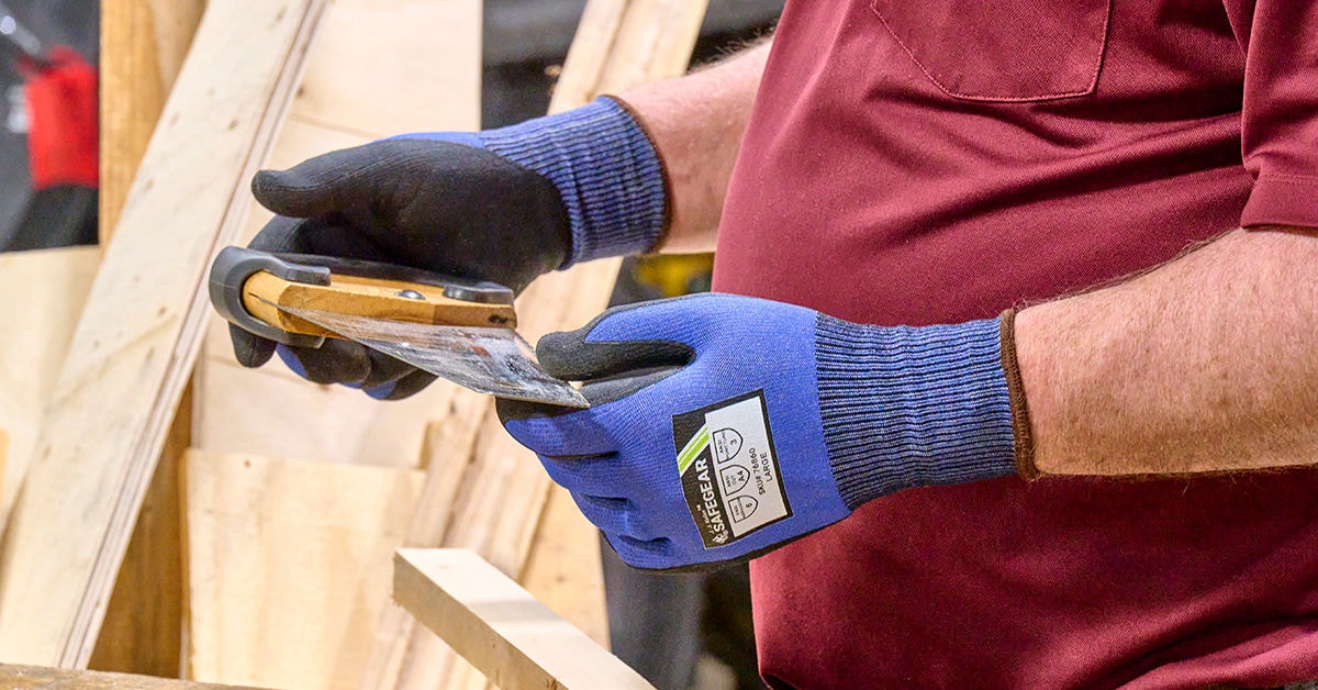 Carpenter wearing J. J. Keller SafeGear protective gloves while smoothing wood, illustrating proper PPE use during a PPE hazard assessment in a workshop setting.