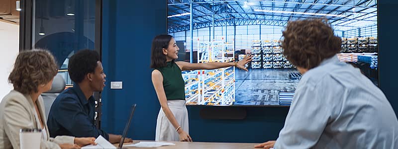 OSHA safety training session with a presenter pointing at a large screen displaying a warehouse layout, attended by four professionals in an office setting.