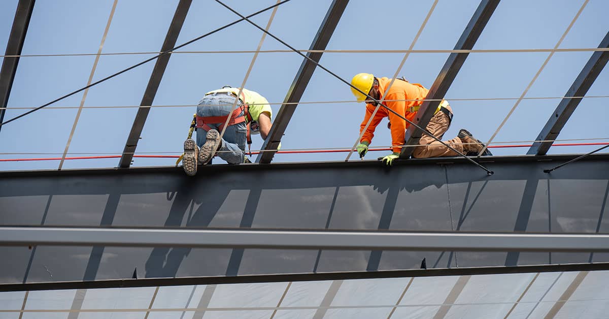 Construction workers wearing fall protection gear while working on elevated steel beam at jobsite.