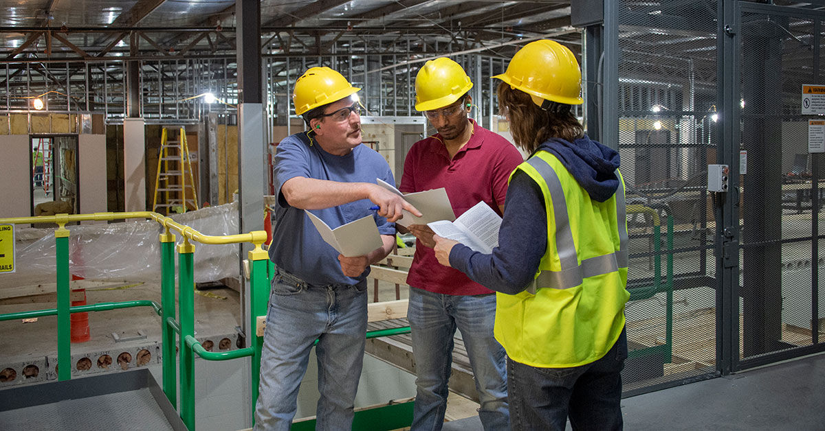 Three workers in safety gear review training documents inside an industrial building under construction.