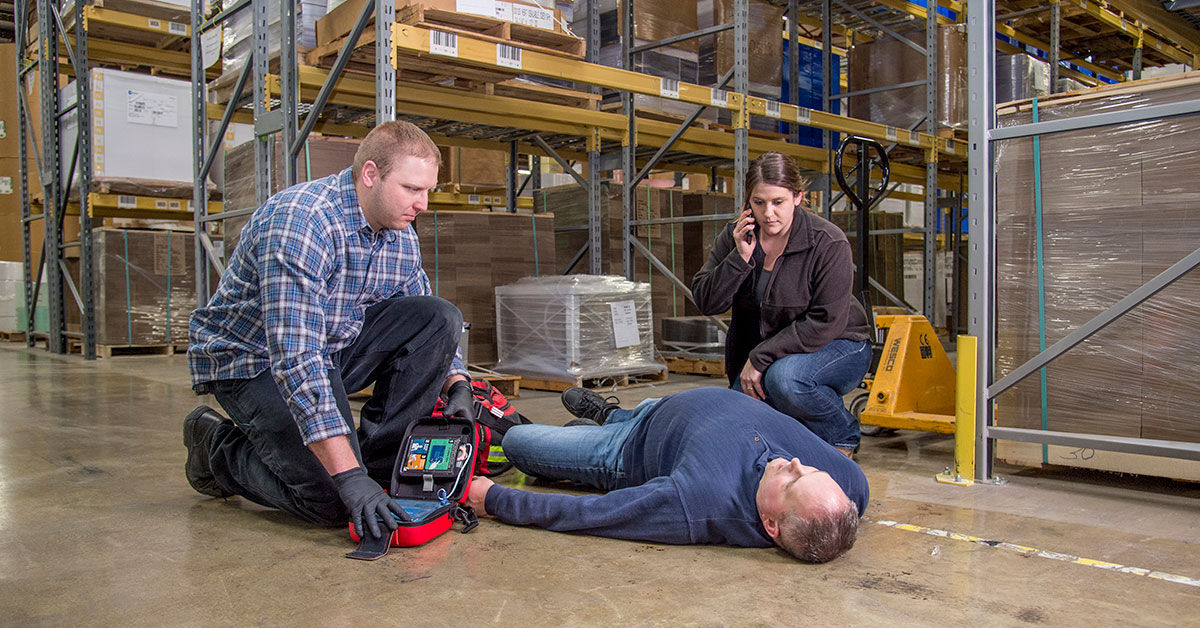 Warehouse emergency response with a worker providing first aid to a collapsed colleague while another calls for help.