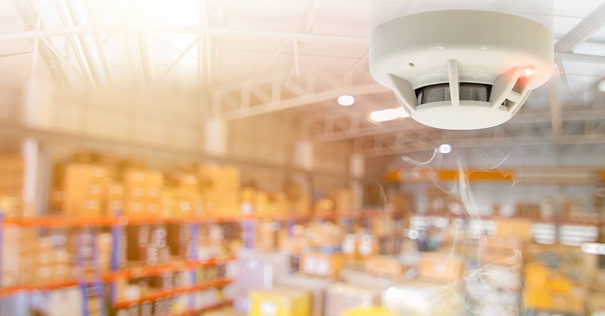 Carbon monoxide detector mounted on the ceiling of a warehouse with shelves of boxes in the background, emphasizing workplace safety and monitoring for CO exposure.