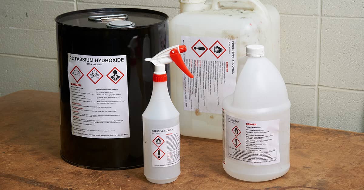 Chemical containers labeled with HazCom-compliant hazard symbols, including potassium hydroxide, isopropyl alcohol, and ethylene glycol.