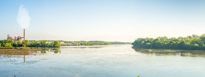 Industrial facility with smokestack near river and green trees under clear sky.