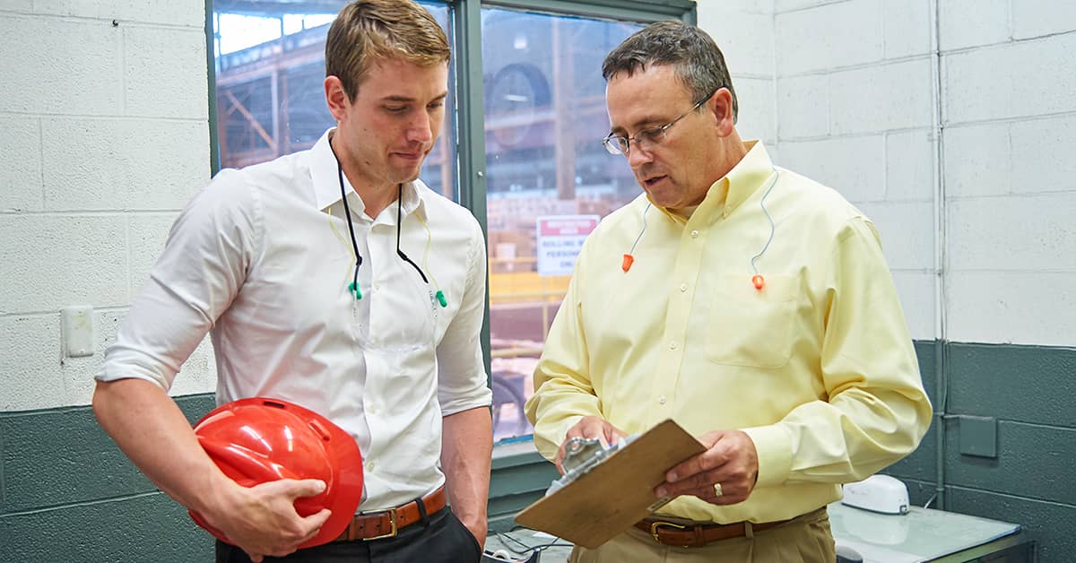 Two safety professionals in a factory setting discussing safety protocols, one holding a red hard hat and the other reviewing notes on a clipboard, both wearing ear protection.