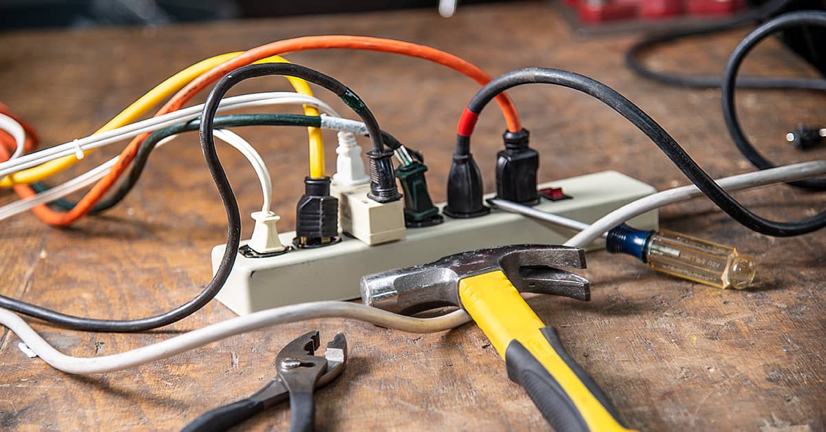 Overloaded power strip with multiple plugs and tools on a wooden workbench, highlighting electrical safety hazards.