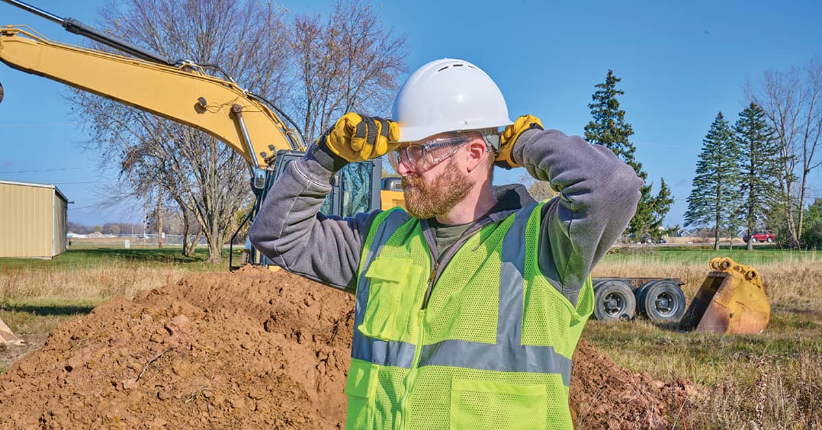 Construction worker adjusting hard hat at excavation site with heavy equipment in the background.