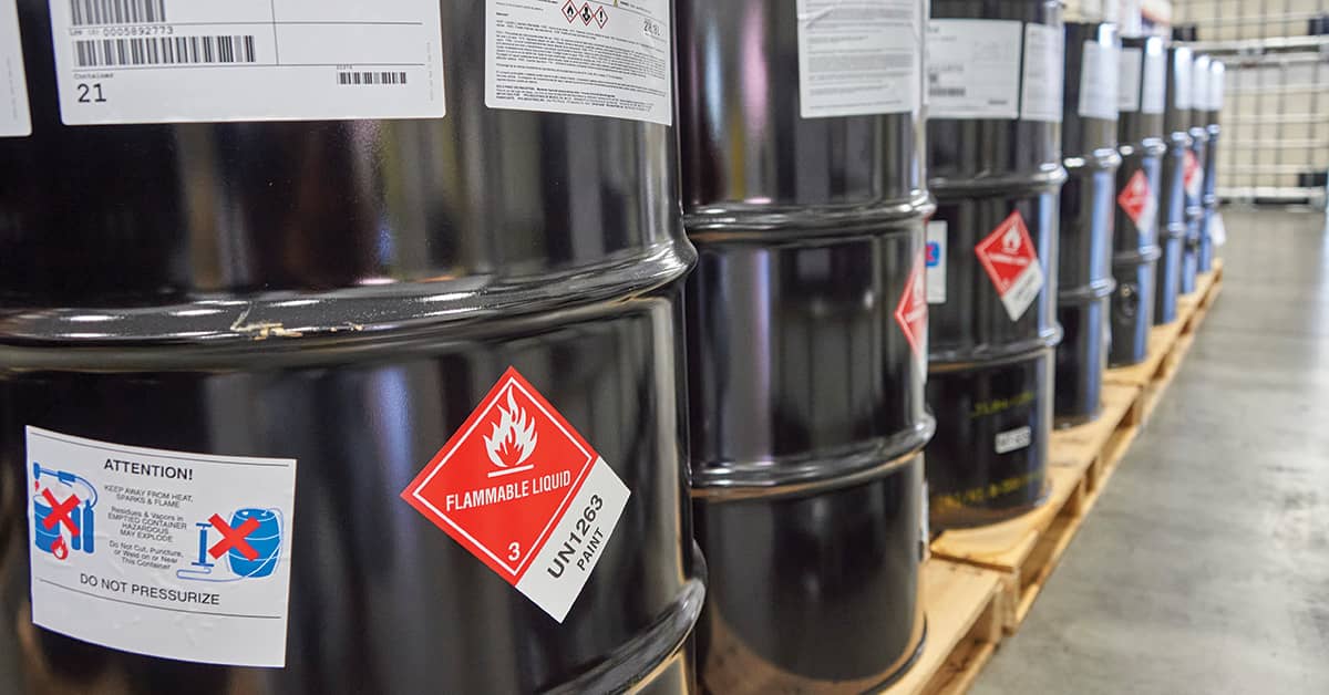 Rows of black metal drums labeled as flammable liquid stored on wooden pallets in a warehouse.