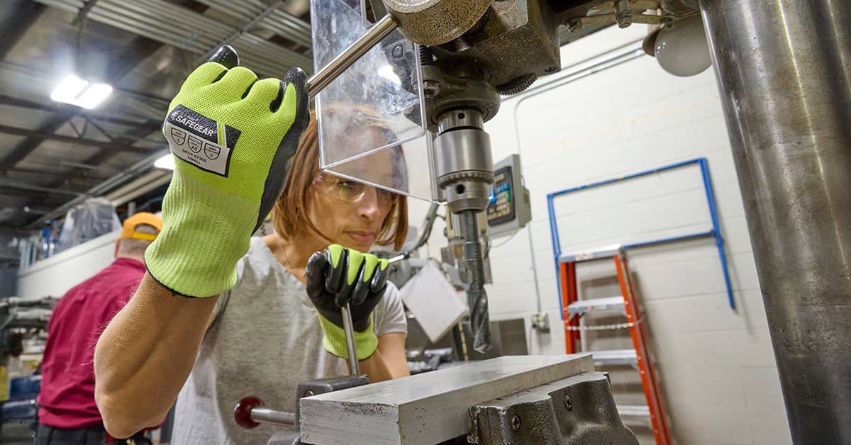 Worker operating a drill press with safety gloves in a workshop, demonstrating machine guarding practices to prevent contact with hazardous moving parts.
