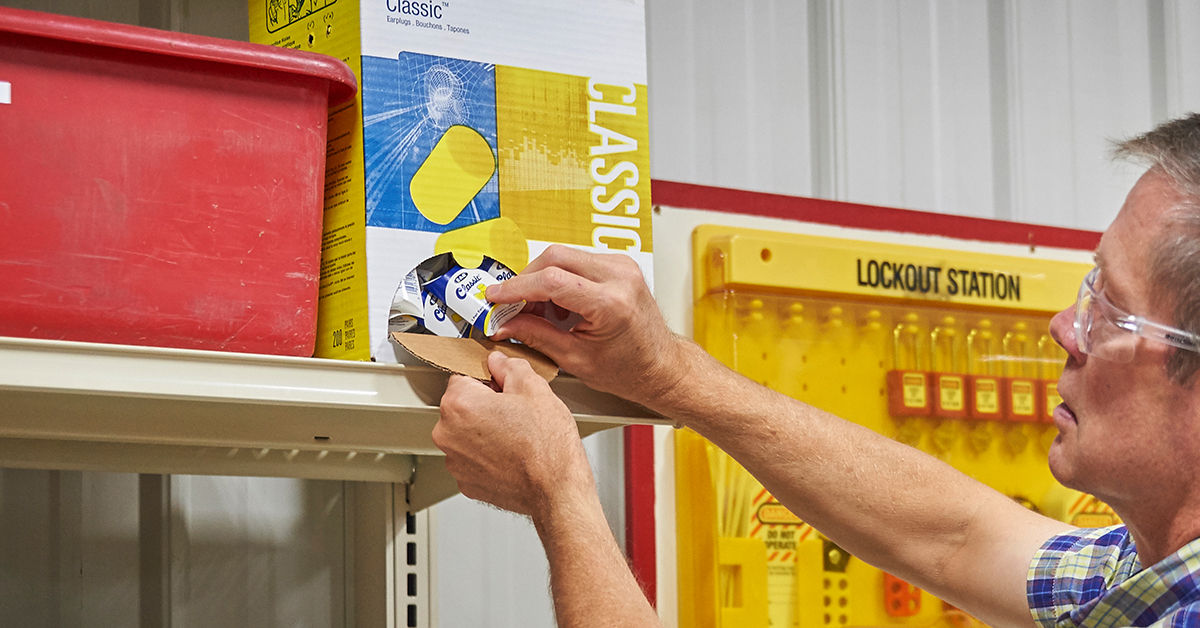 Worker retrieving hearing protection (earplugs) from a dispenser box in an industrial setting, with a lockout station visible in the background.