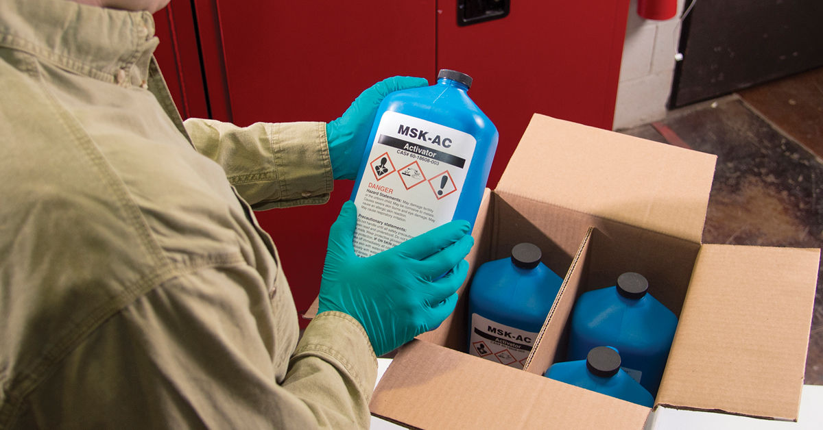 Worker performing hazard communication training task, inspecting labeled chemical containers (MSK-AC) with PPE in an industrial safety setting.