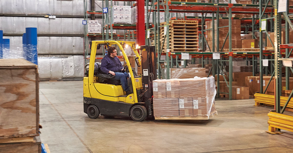 Forklift operator maneuvering a yellow forklift to transport a pallet of goods in a warehouse, surrounded by shelves stocked with boxes and packages.