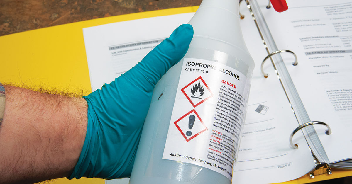 Isopropyl alcohol bottle with OSHA-compliant hazard labels held by gloved hand over workplace safety documents in binder