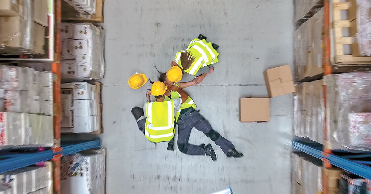 Warehouse workers in high-visibility vests and hard hats assisting an injured colleague on the floor, surrounded by shelves of boxes and packages.