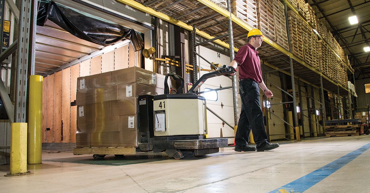 Warehouse worker operating an electric pallet jack to move stacked boxes in a large storage facility with high shelves and organized inventory.