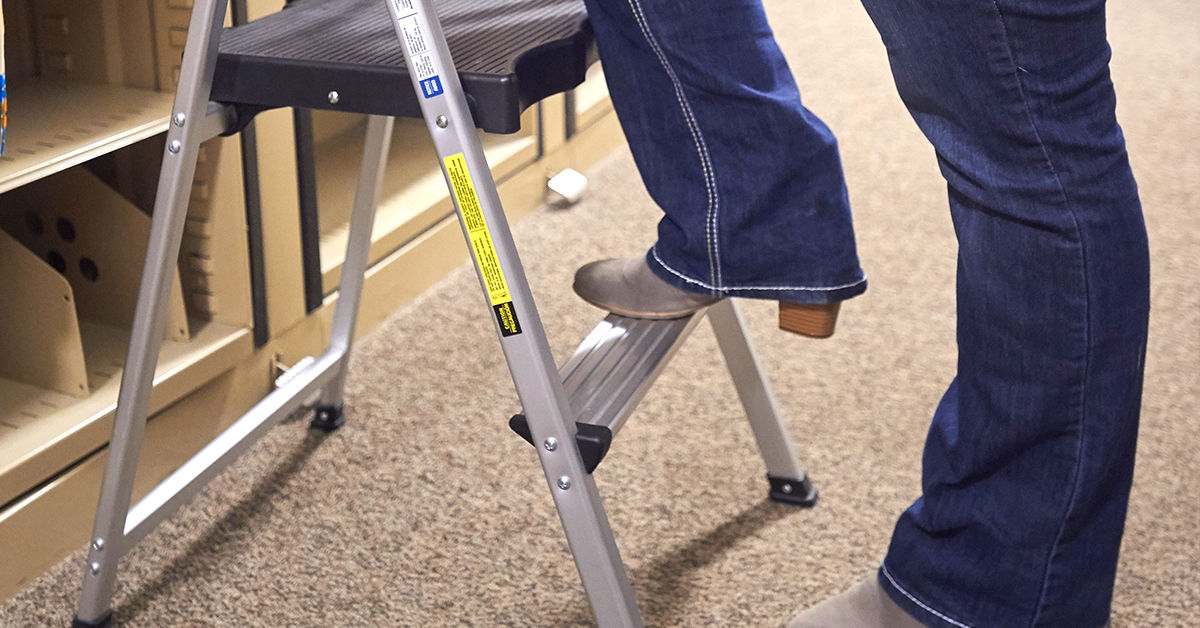 Person wearing jeans and boots stepping onto a small step ladder or step stool in an office or store setting with beige carpet and shelves in the background.