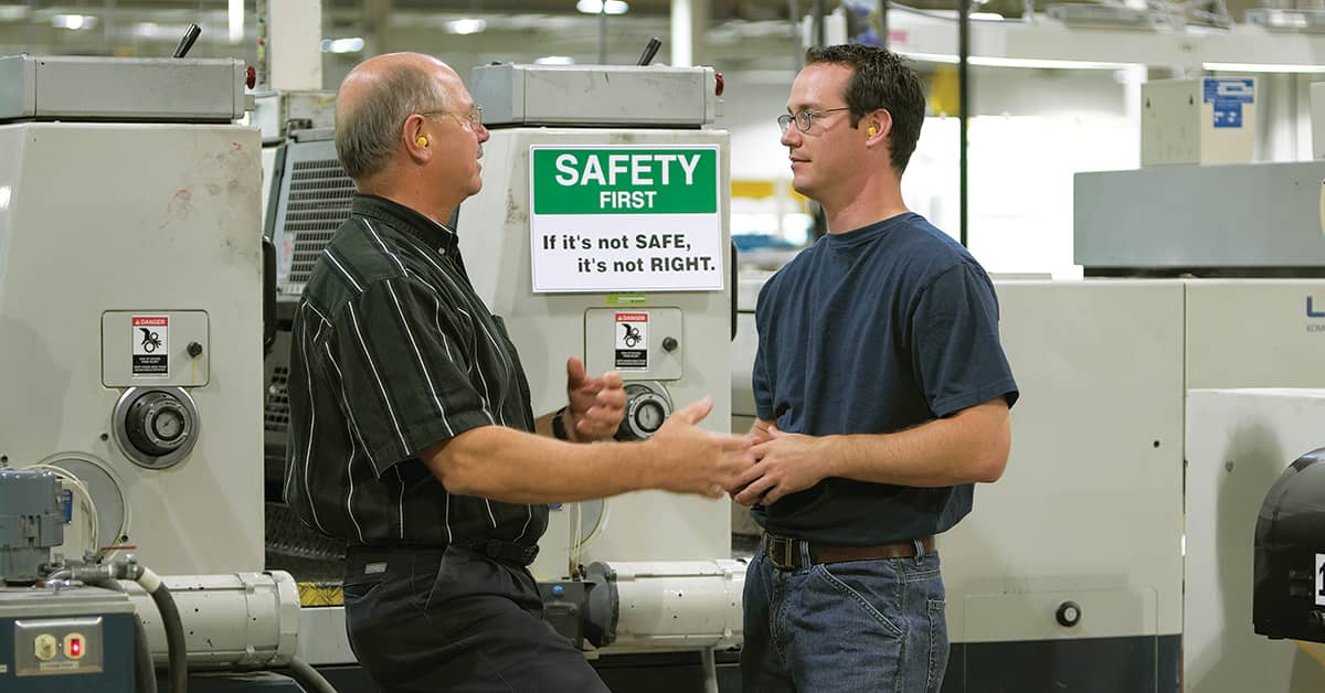 Experienced worker mentoring a new hire on safety procedures in an industrial setting, with a visible 'Safety First' sign—promoting a strong safety culture.