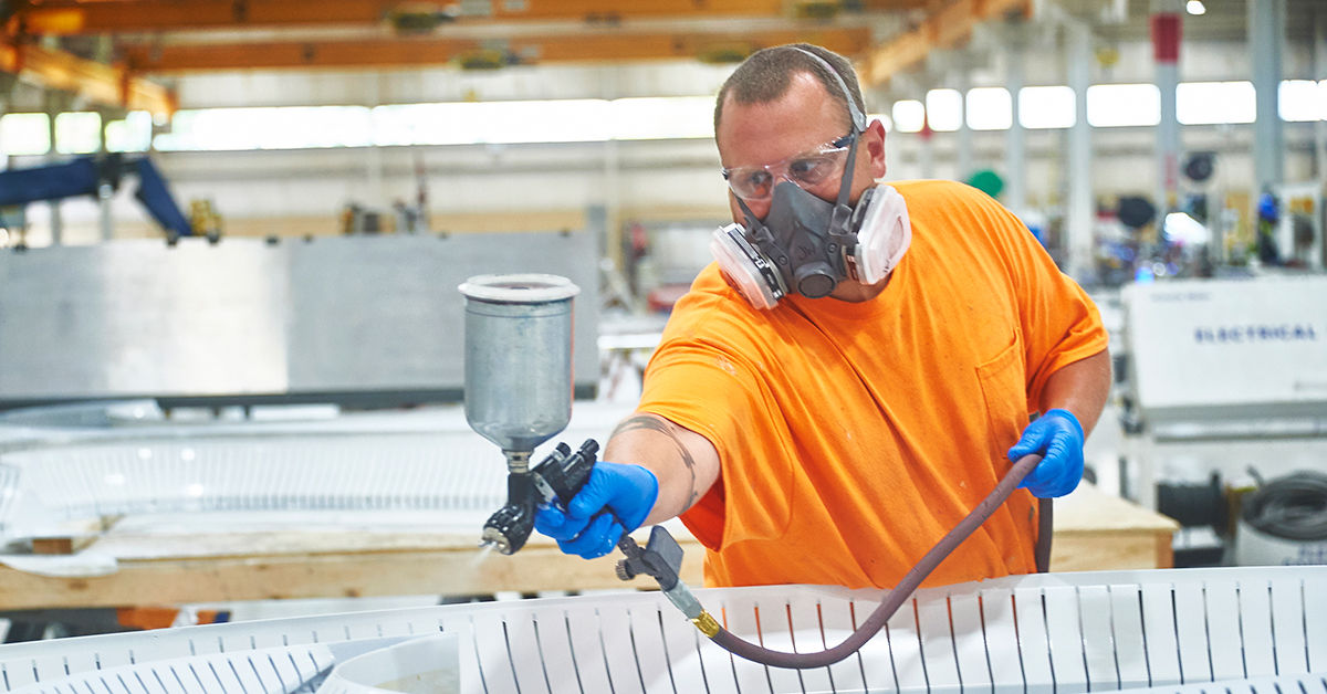 Factory worker in an orange shirt using a spray gun to paint or coat a large industrial component, wearing protective gloves and a respirator mask in a well-lit manufacturing facility.