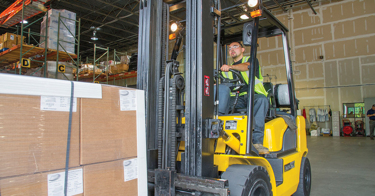 Forklift operator in training moves boxes on pallets, demonstrating real-world skills in a warehouse.