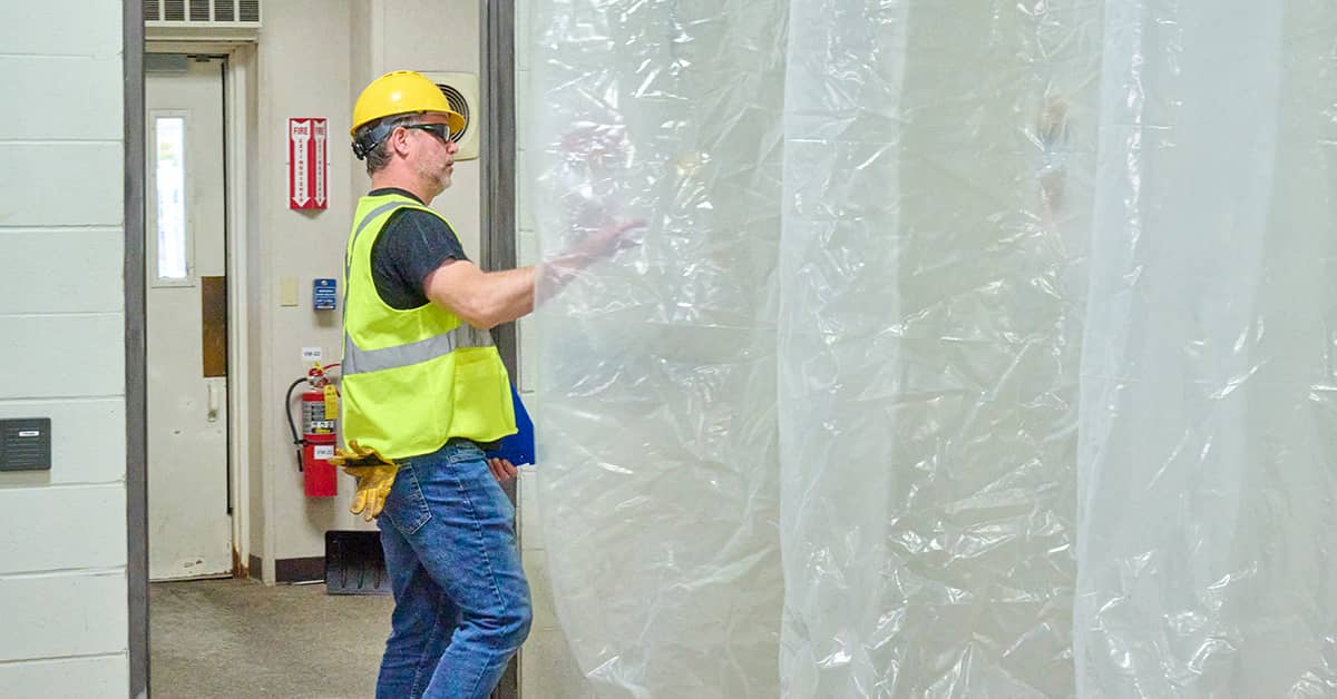 Worker wearing a safety vest and hard hat walking through a plastic dust‑barrier enclosure in an industrial facility.