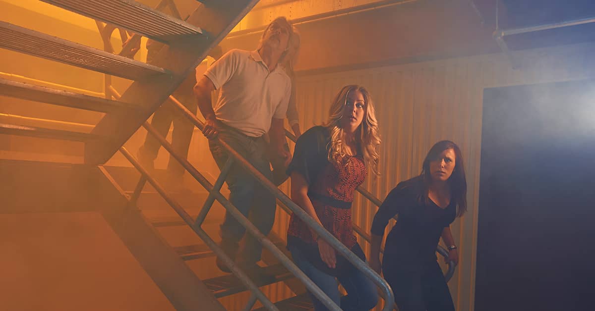 Workers evacuating down a metal staircase in an industrial setting filled with orange smoke, illustrating a fire drill or emergency evacuation scenario.