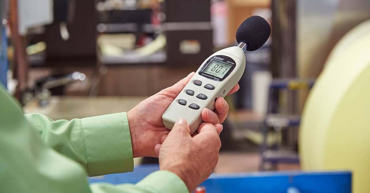 Worker conducting noise monitoring with a digital decibel meter in an industrial setting as part of a hearing conservation program.