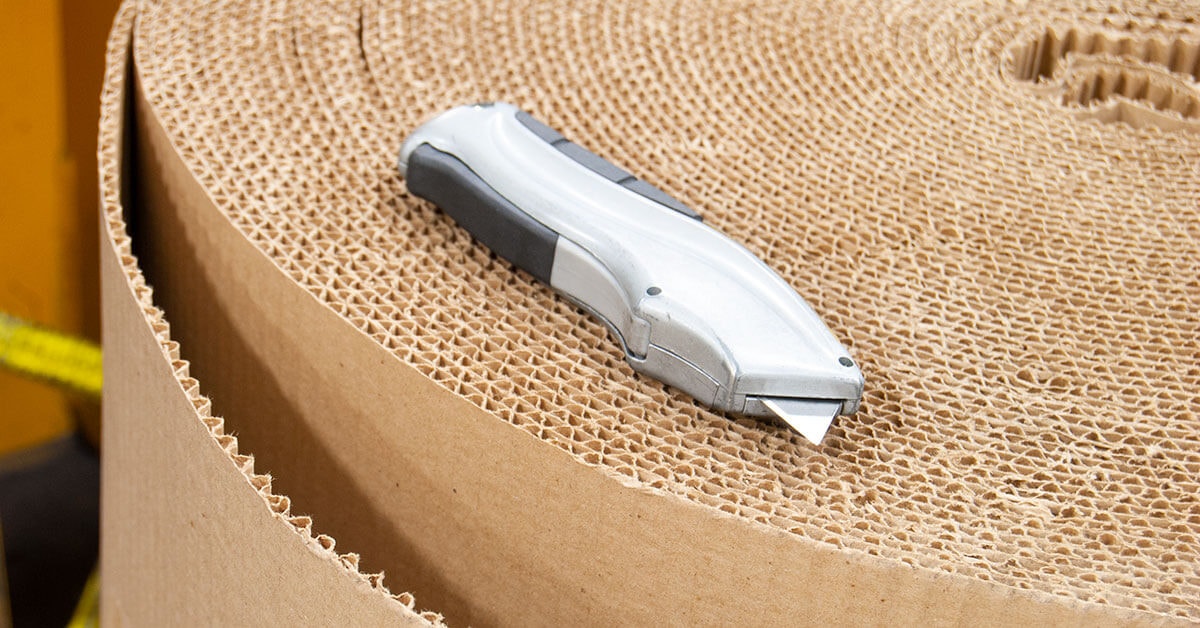 Utility knife resting on a large roll of corrugated cardboard in a packaging warehouse.