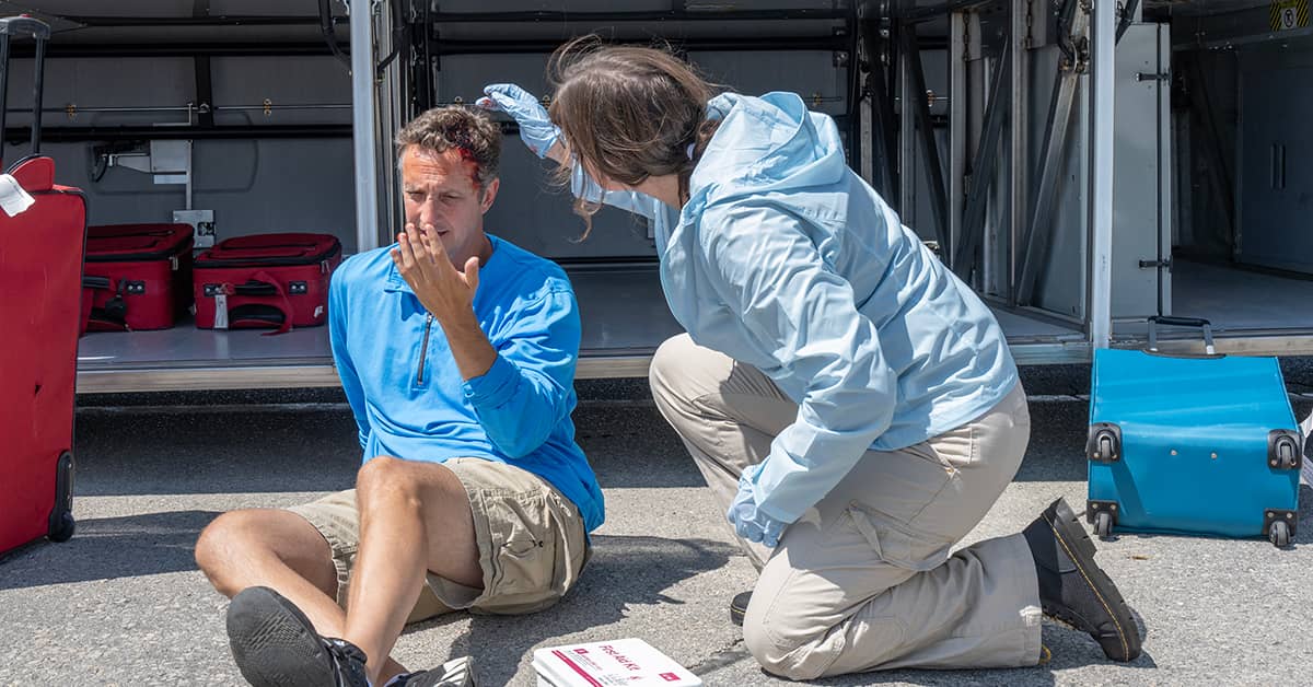 Emergency medical responder providing first aid during a bloodborne pathogen exposure incident, with an injured person near an open ambulance and medical equipment.