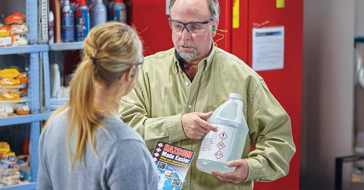 Two employees reviewing hazardous chemical safety procedures, featuring a labeled container with OSHA pictograms and a HazCom safety guide in a workplace setting.