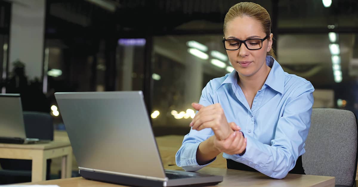 Ergonomic Wrist Injury - Woman at desk in front of computer rubbing her wrist 