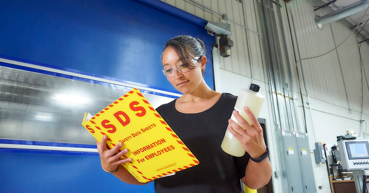 Industrial worker holding Safety Data Sheets (SDS) manual and chemical bottle in facility with equipment and electrical panels.