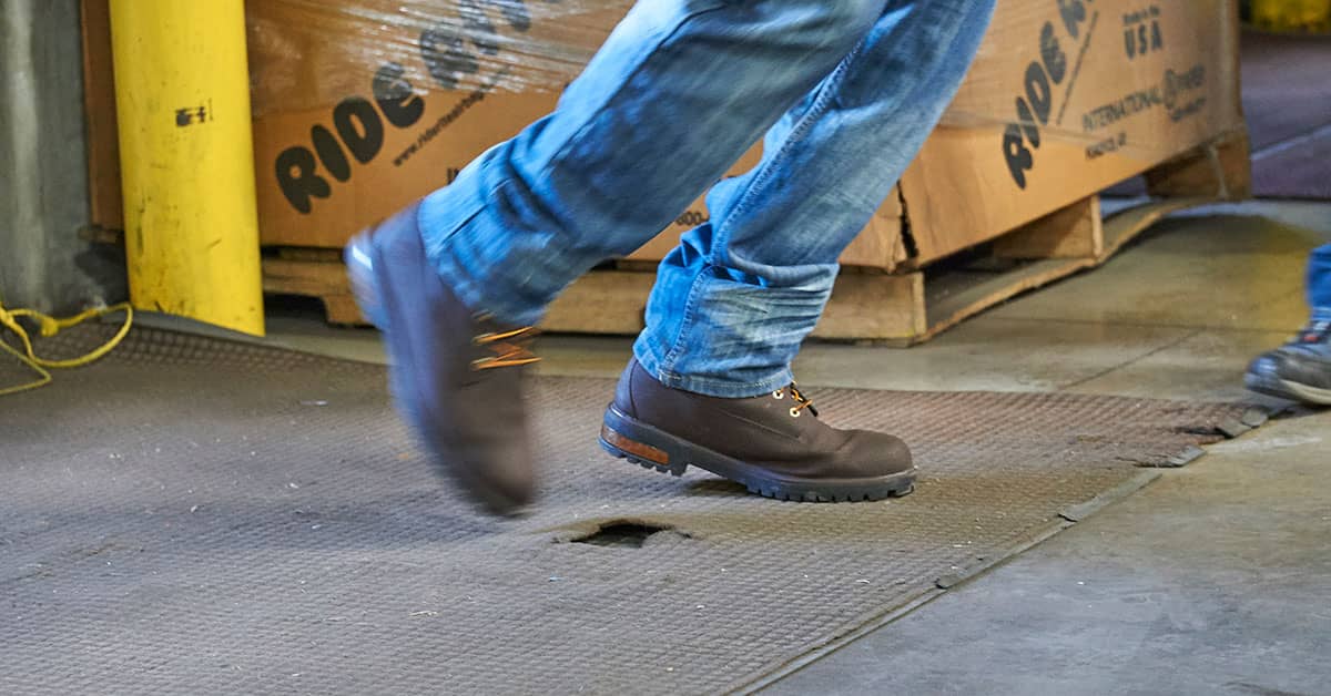 Worker wearing safety boots tripping over a hole in a rug on the warehouse floor, highlighting workplace safety hazards.