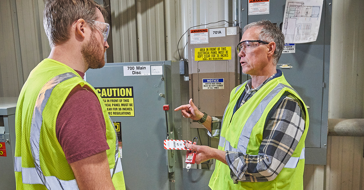 Workers demonstrating lockout tagout training at an electrical panel with visible safety vests, caution signs, and warning labels.