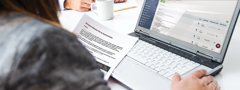 Woman holding Emergency Response Safety Plan from J. J. Keller SAFFETY MANAGEMENT SUITE, the plans & policies section of the site shows on a laptop screen.